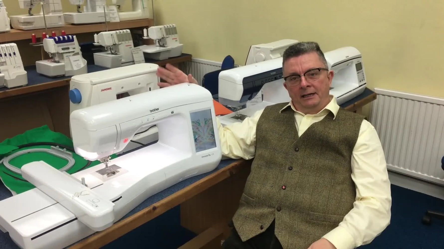 A presenter sits beside a Brother Innov-is V3 embroidery machine in a sewing machine shop, introducing the machine and its large-format capability.