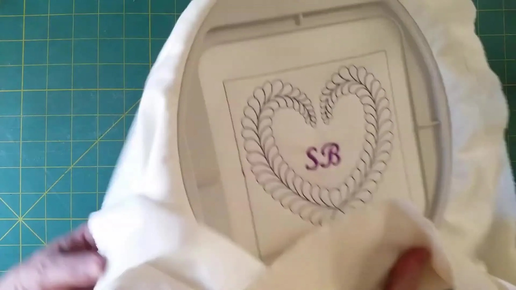 The finished quilt block is held up to the camera, showing centered purple initials inside a feathered heart with a professional, clean-looking result.