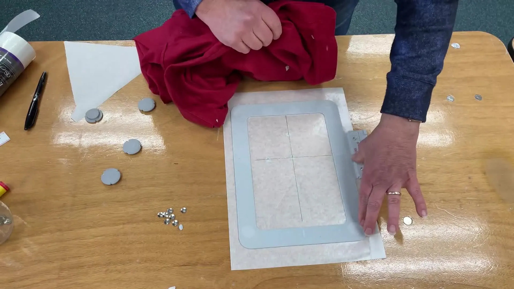 A red button-down shirt is being aligned onto a sticky stabilizer surface using a center thumbtack as a precise registration point inside a magnetic hoop.