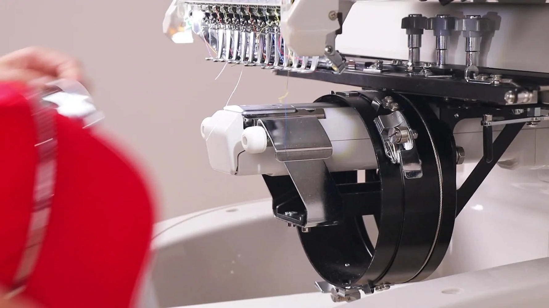 A hooped red cap in a metal cap frame is held in front of the machine driver, ready to be mounted for cap embroidery.